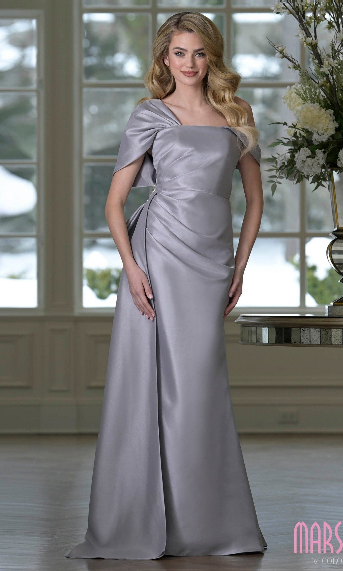 A woman stands indoors in front of large windows and a floral arrangement, wearing the Marsoni MV1405 long formal dress in grey, featuring an asymmetric neckline and draping details.